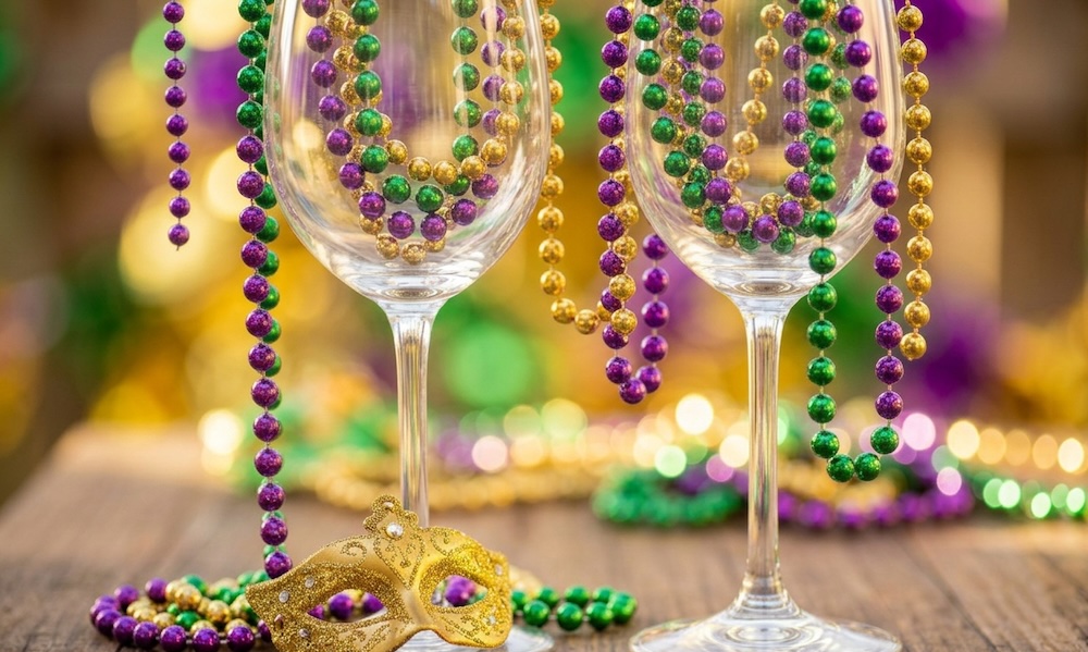 Purple, green, and gold Mardi Gras beads draped over wine glasses with a gold mask